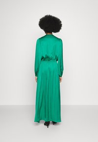 Grön satäng maxiklänning med långa ärmar, rynkad midja, slät textur och en flödande silhuett. Bakvyn visar elegant drapering.