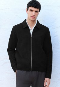 Melns rāvējslēdzēja džemperis ar strukturētu rakstu, ar apkakli un garām piedurknēm, valkāts virs gaišas krāsas krekla, kombinēts ar pelēkām biksēm.