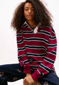 Bordowa koszulka polo z długim rękawem w paski w kolorze białym i granatowym, z białym kołnierzykiem oraz ozdobnymi haftami na ciemnych spodniach dżinsowych.