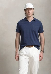 Polo shirt bleu marine à manches courtes avec une poche poitrine, associé à un pantalon plissé blanc et une casquette blanche. Ceinture brune et montre visibles.