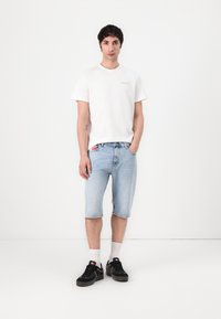 Weißes Baumwoll-T-Shirt, blaue Jeansshorts mit einem roten Etikett, graue Turnschuhe und weiße Socken, die vor einem schlichten weißen Hintergrund stehen.