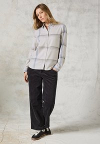 Camicia button-up grigia chiara con motivo a quadri blu e grigio, abbinata a pantaloni a gamba larga in corduroy neri e sneaker nere. Sfondo testurizzato.