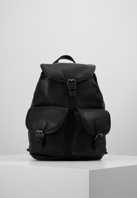 Czarny skórzany plecak z górną klapą i zapięciem, z dwiema przednimi kieszeniami z zapięciami, umieszczony na białym piedestale na jednolitym tle.