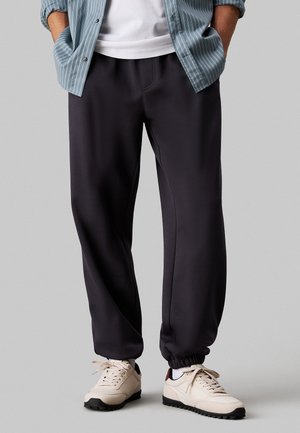 Ciemnoszare dresy wykonane z gładkiego materiału, z elastycznym pasem i ściągaczami, w połączeniu z jasnymi butami sportowymi.