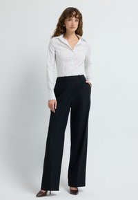 Witte blouse met zwarte verticale strepen, gecombineerd met hoge zwarte broeken, beide op maat gemaakt voor een nette fit en silhouet.