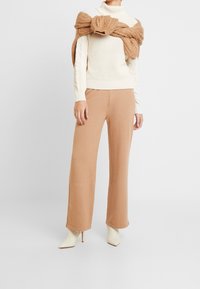 Pull col roulé en tricot torsadé crème, pantalon large beige, et bottines à talons beiges, avec une étole en tricot brun texturé drapée sur les épaules.