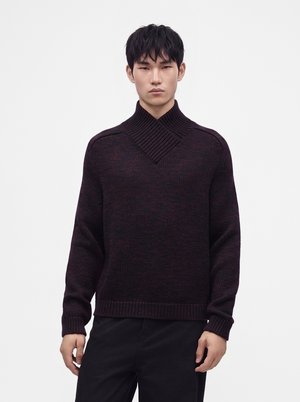 Jeune homme portant un pull en laine foncé avec un large col montant côtelé et un pantalon noir, debout devant un fond blanc uni.