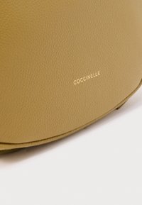 Gele leren handtas met een gladde textuur en een afgeronde vorm. De merknaam "Coccinelle" is in goud gegraveerd.