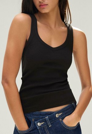 Fiatal nő fekete ujjatlan bordázott felsőben és kék farmerben, kezét zsebében tartva, semleges háttér.