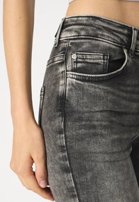 Jean noir délavé avec une coupe ajustée, présentant des coutures visibles, une poche avant et une texture usée sur le côté.