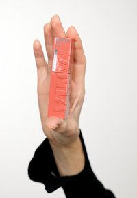 Rossetto in tubetto rettangolare rosso etichettato "Superstay Vinyl Ink" con lettering in rilievo, tenuto verticalmente tra le dita su uno sfondo chiaro.