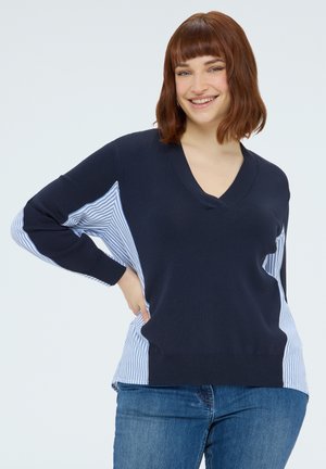 Jeune femme aux cheveux bruns mi-longs souriante, portant un pull bleu marine à col en V avec des panneaux latéraux rayés bleu et blanc, et un jean bleu.