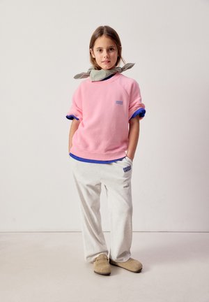 Ragazza giovane in piedi con le mani nelle tasche, indossa una felpa rosa, una maglietta blu sotto, pantaloni della tuta chiari, scarpe beige e una sciarpa legata intorno al collo.