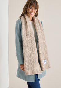 Bufanda de punto beige con un patrón de espiga, hecha de material suave, drapeada alrededor del cuello. Etiqueta con la marca visible en un extremo.