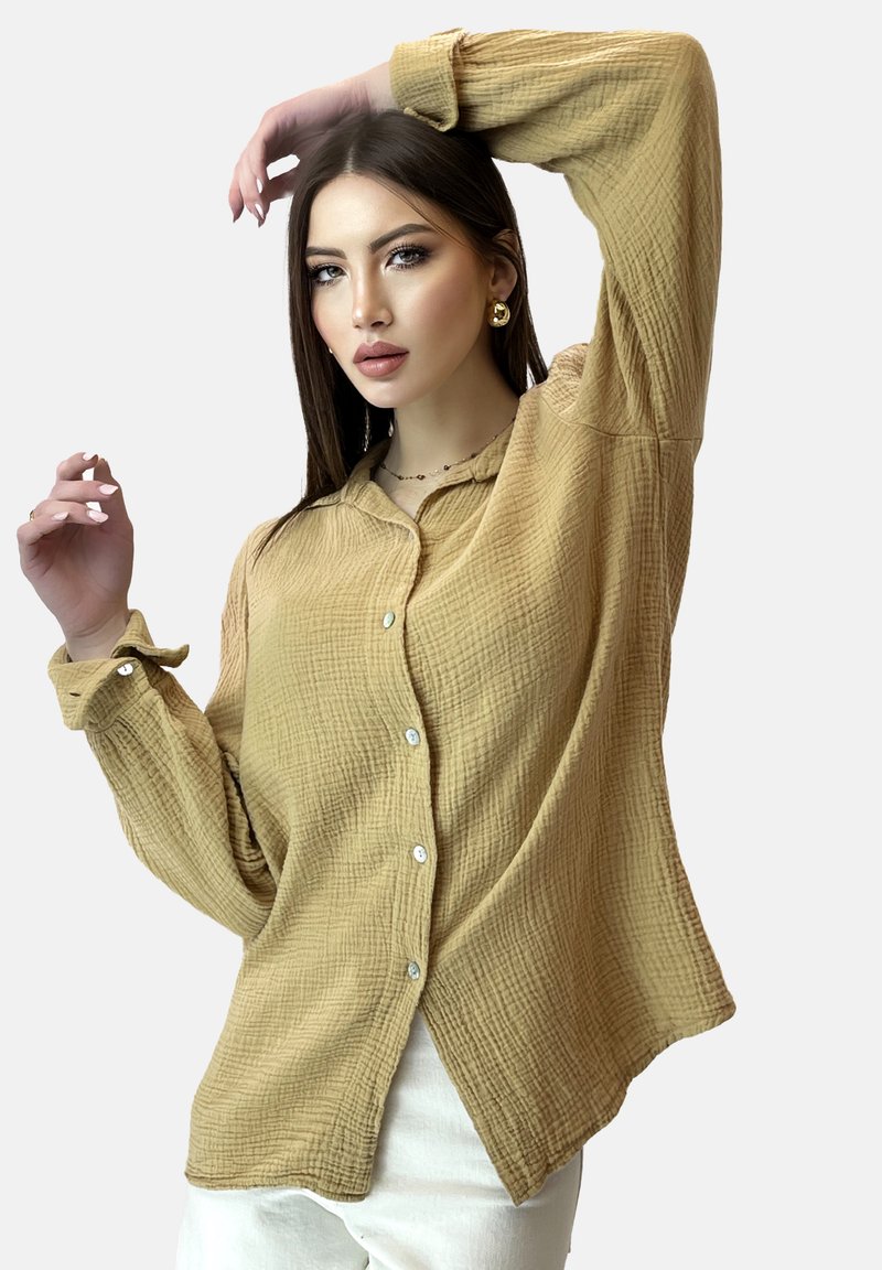Mujer con el cabello largo y oscuro, vistiendo pendientes de aro dorados y una camisa amarilla mostaza de botones, posando con un brazo levantado.