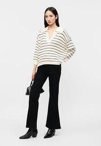 Kobieta ubrana w biało-czarny sweter w paski, czarne rozszerzane spodnie, czarne botki i trzymająca małą czarną torebkę na jednolitym tle.