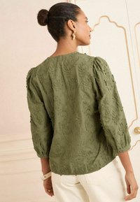 Blouse brodée vert olive avec des manches trois-quarts, présentant des motifs floraux et une finition texturée, complétée par un col rond.