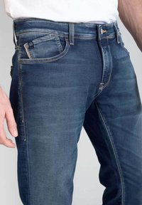 Vaqueros de denim azul oscuro con un corte ajustado, que cuentan con un diseño de cinco bolsillos, costuras en contraste y un cierre de cremallera con un botón metálico.