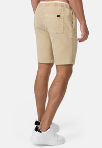 Beige Cordshorts mit einem elastischen Bund und einer Gesäßtasche. Glatte Textur und Oberschenkellänge, kombiniert mit weißen Sneakers.