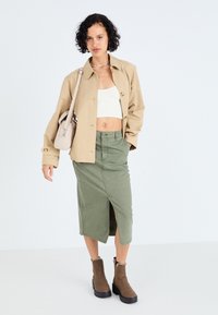 Jachetă oversized bej, top alb scurt, fustă midi verde cu o crăpătură, cizme maro până la gleznă cu o talpă neagră groasă și o geantă mică bej.