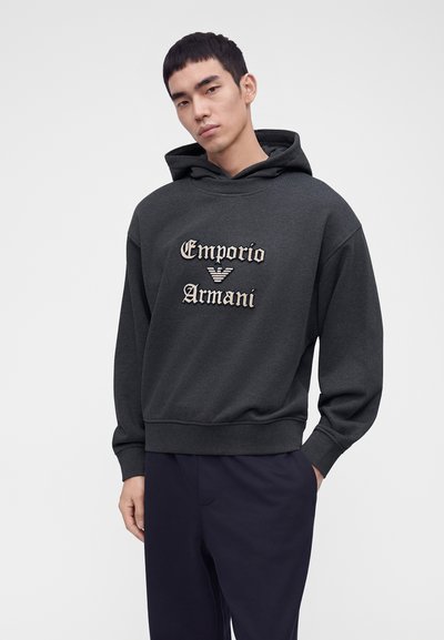 Homme portant un sweat à capuche Emporio Armani gris foncé et un pantalon bleu marine, debout avec une main dans la poche sur un fond uni.