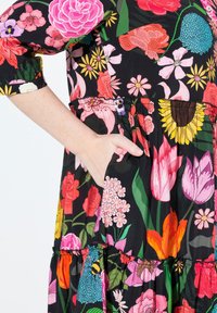 Kwiatowa sukienka z czarnego materiału, z żywymi różowymi, żółtymi i zielonymi wzorami; ma marszczoną talię i boczne kieszenie.