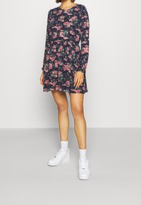 Kukkakuvioinen pitkähiha-mekko laivastonsinisellä pohjalla, jossa vaaleanpunaisia ja vihreitä kuvioita, vartalonmyötäinen yläosa, liehukehelmainen hame, yhdistetty valkoisiin tennareihin ja nilkkasukkiin.