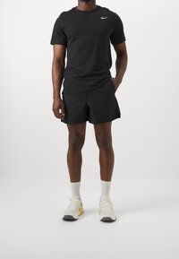 Camiseta negra de manga corta de Nike con logo blanco, combinada con pantalones cortos negros con textura. Calcetines deportivos blancos y zapatillas multicolores completan el conjunto.