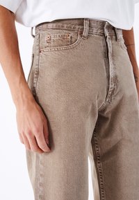 Pantalones vaqueros de mezclilla beige con cintura alta, remaches de cobre, diseño de cinco bolsillos y una textura ligeramente descolorida para un aspecto casual.