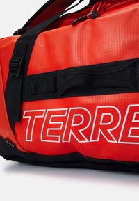 Röd duffelväska med en texturerad yta, med svarta remmar och en djärv vit text "TERRE" längs sidan. Inkluderar dragkedja och detaljer i hårdvara.