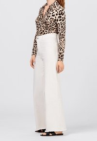 Donna che indossa un top a maniche lunghe leopardato beige, pantaloni bianchi a vita alta e larga, e sandali neri con apertura sulle dita, in posizione laterale.