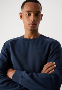 Ung mann som har på seg en marineblå Marc O'Polo hettegenser med armene krysset, mot en enkel lys bakgrunn.