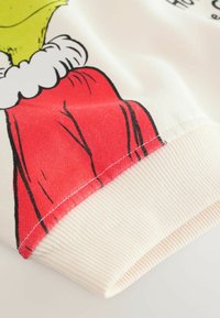 Biely textil s rozprávkovým Grinchem v červenom plášti. Manžeta je rebrovaná a prírodná, s červenými detailmi a čiernymi obrysmi.