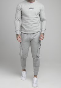 Sudadera gris y pantalones de chándal hechos de un tejido suave, con un logo en la sudadera y bolsillos tipo cargo en los pantalones. Combinados con zapatillas blancas.
