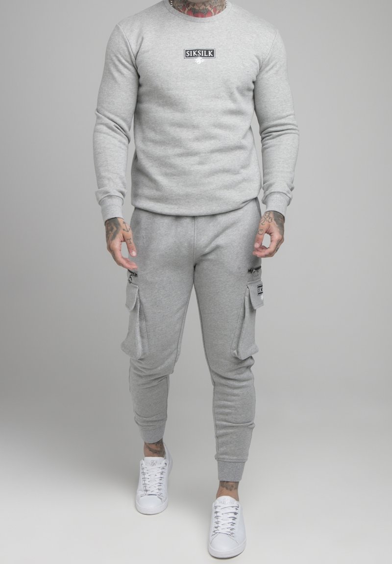 Sudadera gris y pantalones de chándal hechos de un tejido suave, con un logo en la sudadera y bolsillos tipo cargo en los pantalones. Combinados con zapatillas blancas.