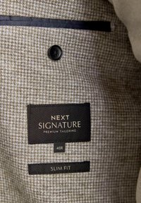Braun kariertes Innenfutter mit schwarzem Knopf, versehen mit Etiketten: "NEXT SIGNATURE", "PREMIUM TAILORING", "40R" und "SLIM FIT."