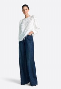 Sieviete valkā balti teksturētu garroku jaciņu un tumši zilas platām kāju bikses, stāvot ar vienu roku kabatā pret vienveidīgu fonu.