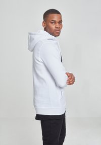 Hvit hettegenser med en strømlinjeformet design, mykt stoff og kengurulomme, kombinert med svarte bukser, vist fra siden.