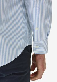 Vaaleansininen ja valkoinen raidallinen paita, jossa on pyöreä helma. Varustettu nappihihoilla ja sileällä kankaan tekstuurilla. Paita on puettu tummien housujen päälle.