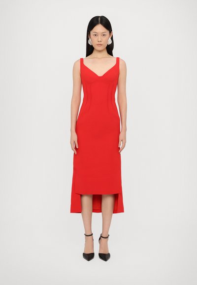 Red fitted dress with a V-neck, thick straps, and structured bodice. Features high-low hem and smooth fabric. Paired with black ankle strap heels.