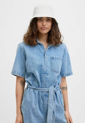 Vrouw met krullend bruin haar draagt een witte bucket hat en een lichtblauwe denim jumpsuit met korte mouwen en een riem, met tatoeages op beide onderarmen.