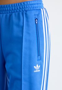 Modre športne hlače z belimi poudarnimi črtami. Imajo žep s zadrgo in logo Adidas v beli barvi na spodnji levi strani.