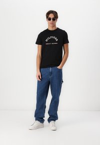 T-shirt en coton noir avec logo blanc "HILFIGER EST. 1985", associé à un jean en denim bleu délavé et des baskets blanches. Modèle portant des lunettes de soleil.