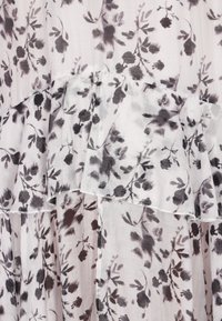 Leichtes Kleid aus Stoff mit einem geschichteten Design und einem schwarzen Blumenmuster auf einem sanften weißen Hintergrund. Die Ränder sind mit dezenten Nähten versehen.