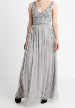 Mouwloze zilveren jurk met geborduurde bloemen op het lijfje, diepe V-hals en vloeiende tule rok, gecombineerd met zwarte schoenen.