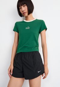 Žalias medvilninis marškinėliai su balta apykakle ir išsiuvinėtu "nike" logotipu; derinami su juodais šortais, turinčiais elastingą juosmenį ir logotipo akcentą.