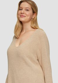 Beige gebreide trui met een V-hals, lange mouwen en subtiele zijdetails, gemaakt van een zachte, gestructureerde stof.