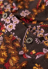 Tissu à motifs avec une base brun foncé ornée de motifs floraux en rose, rouge et jaune. Comprend un bouton rond noir à quatre trous.