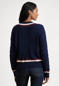 Laivastonsininen palmikkoneuleinen neulepaita, jossa on resorit hihansuissa ja helmassa; kauluksella ja alaosassa viininpunaiset ja valkoiset raidalliset korostukset.