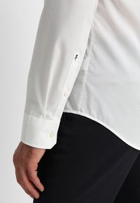 Camicia bianca con polsini a bottone, con un sottile dettaglio ricamato nero. Tessuto morbido e vestibilità sagomata abbinate a pantaloni scuri.